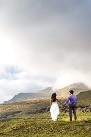 Couple de mariés en tenue de cérémonie dans un paysage vallonné, sous un ciel nuageux.